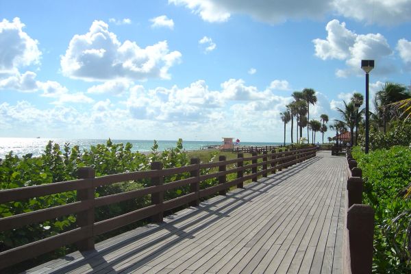 A wooden boardwalk runs parallel to a beach with the sea on the left, lush greenery, and palm trees, under a sky with scattered clouds.