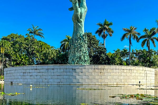 A large hand sculpture emerges from a stone base, surrounded by a reflective pond with water lilies, against a backdrop of trees and blue sky.