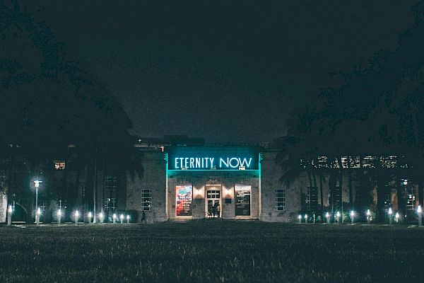 A building is lit up at night with the words "Eternity Now" prominently displayed above its entrance, surrounded by trees and pathway lights.