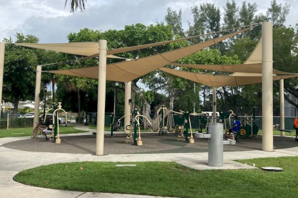 An outdoor exercise area with fitness equipment, shaded by fabric canopies, surrounded by green grass and walkways, under a partly cloudy sky.