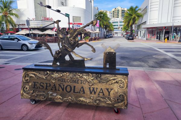 A street scene depicting a "Welcome to Espanola Way" sign with a decorative sculpture on top, palm trees, cars, and buildings in the background.