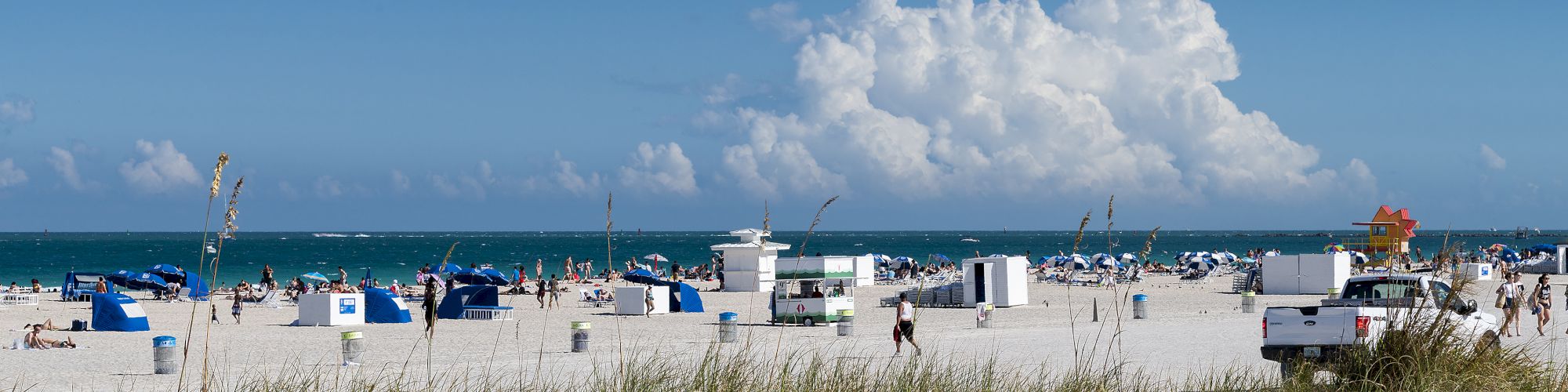 A beach scene with people, blue cabanas, beach chairs, and a lifeguard stand, viewed from behind grass dunes with blue sky and clouds in the background.