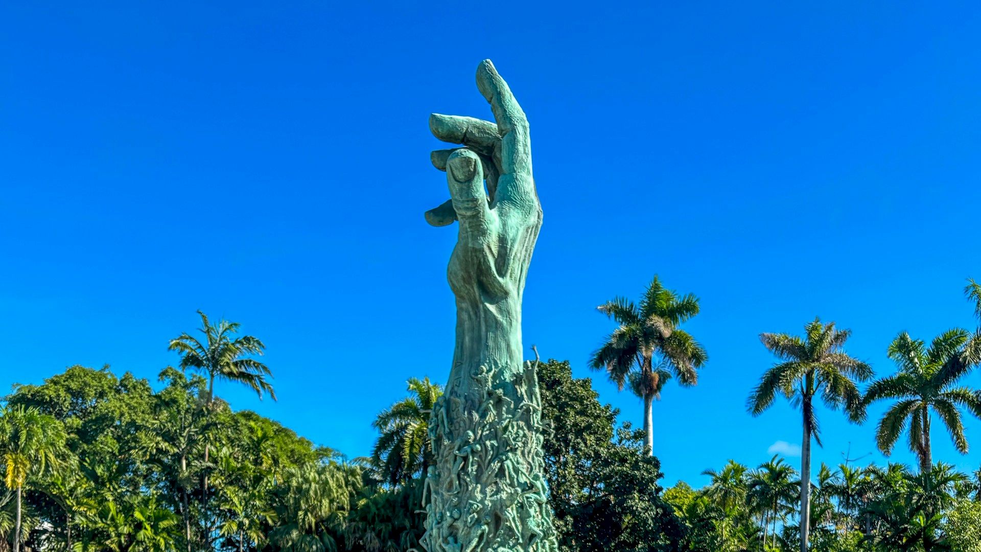 A large sculpture of a hand emerges from the center of a pool surrounded by palm trees and greenery, with water lilies in the foreground.