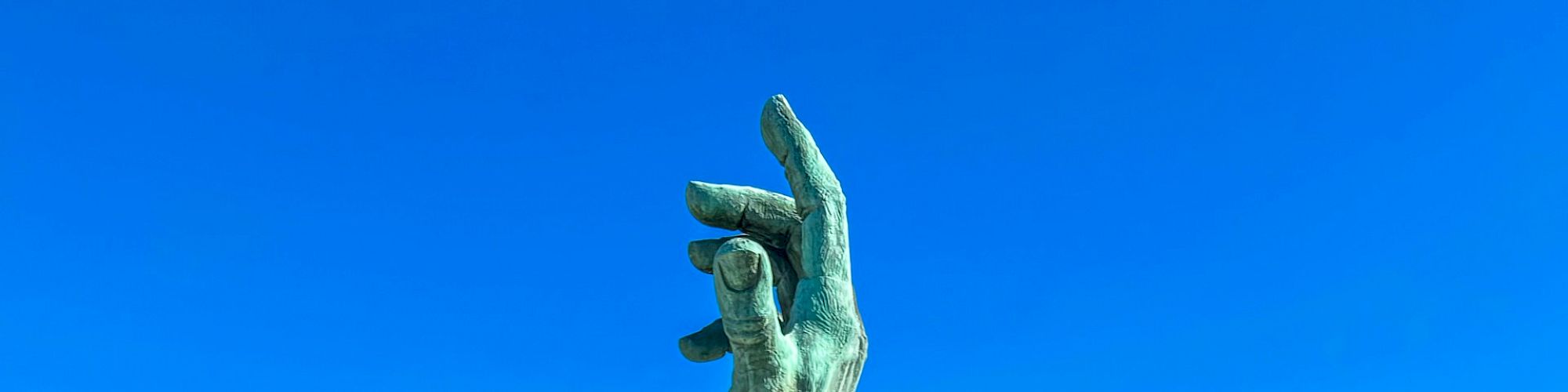 A large sculpture of a hand emerges from the center of a pool surrounded by palm trees and greenery, with water lilies in the foreground.