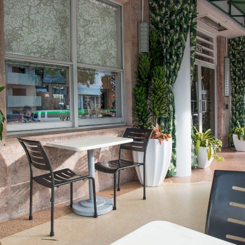 An outdoor café area with tables, chairs, and large potted plants, featuring a shaded sidewalk and a leafy decor theme throughout the space.