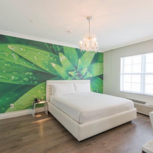 A bright bedroom with a large bed, green leaf mural on the wall, chandelier, window, and chair near a wooden floor.