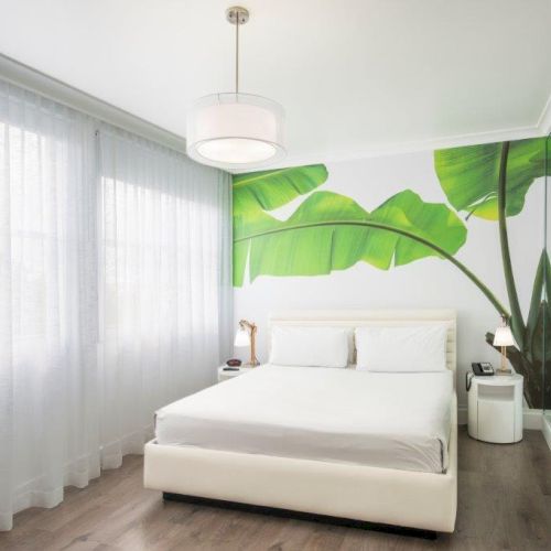 A modern bedroom with a white bed, tropical leaf wall art, mirrored closet doors, wooden flooring, large sheer curtains, and a ceiling light fixture.