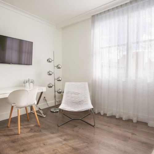 A modern, minimalist room with a wall-mounted TV, desk and chair, unique floor lamp, and a white chair by a window with sheer curtains.