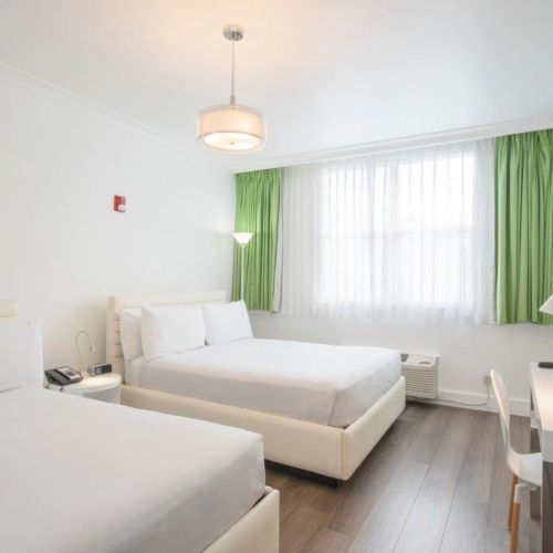 A hotel room with two double beds, white linens, green curtains, a wall-mounted TV, desk, lamp, wooden floor, nightstand, and modern lighting ends the sentence.