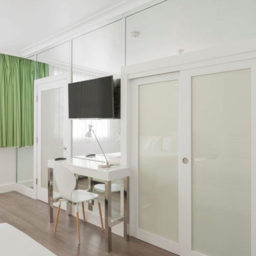 A modern, white-themed bedroom with green curtains, a TV, a desk and chair, a frosted glass sliding door, and a window letting in natural light.