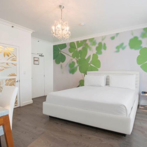 A modern bedroom with a white bed, green leafy wall mural, chandelier, desk, and chair, featuring a clean design and wooden floor.