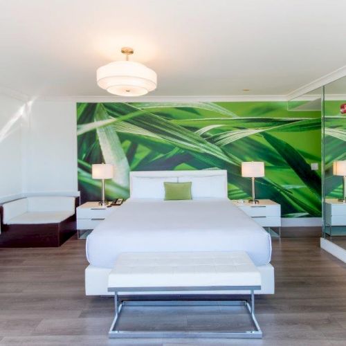 A modern bedroom with a large bed, green plant-themed wall art, side tables, lamps, a bench, mirrored wall, and wooden flooring.