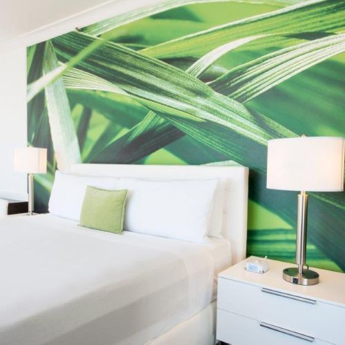 A modern bedroom featuring a king-size bed with white linens, a green pillow, bedside tables with lamps, and a mural of green leaves on the wall.