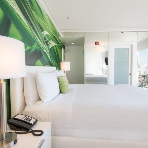 A modern bedroom with a nature-themed wall mural, a white bed, bedside lamp, phone, and reflective closet doors. A window shows city skyscrapers.