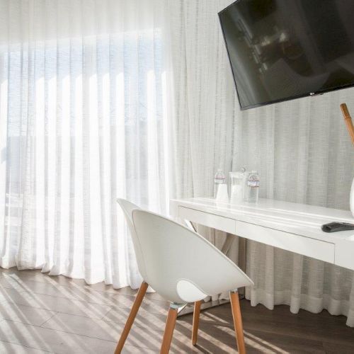 A modern room with a white desk, chair, a TV mounted on the wall, white curtains, and a vase with wooden sticks on the desk.