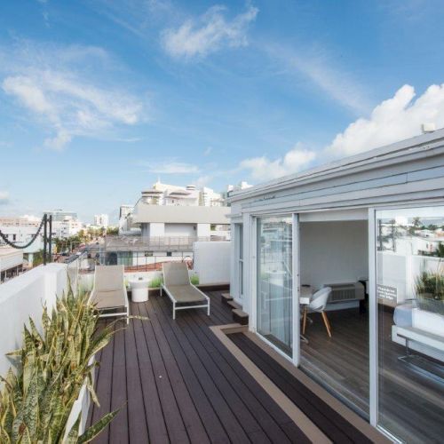 The image shows a rooftop deck with lounge chairs, potted plants, a seating area, and a partially open glass door offering a cityscape view under a blue sky.