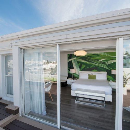 A modern bedroom with a green accent wall and a large bed, viewed through sliding glass doors, leads to a rooftop deck with lounge chairs.