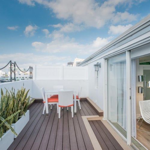 The image shows a rooftop patio with a small table and chairs, potted plants, and an indoor lounge area visible through sliding glass doors.