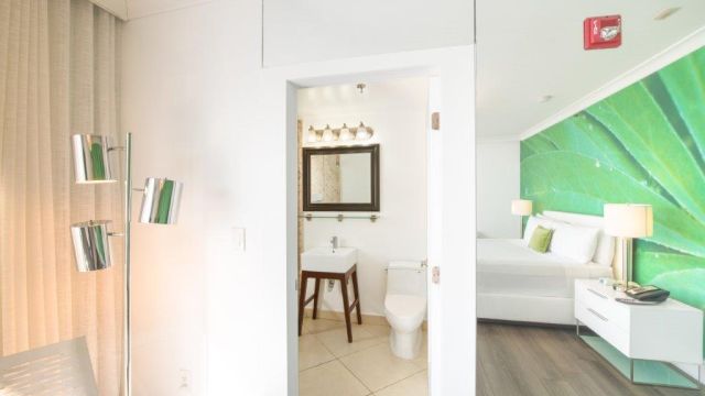 A modern hotel room with a green accent wall, white bed, and bathroom in the background.