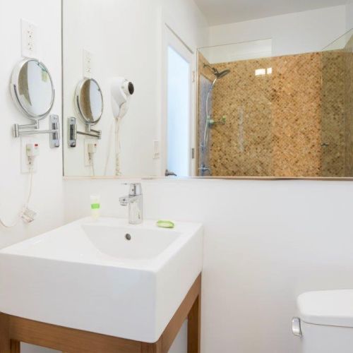 A modern bathroom features a sink, toilet, wall-mounted hairdryer, mirror, and a shower with a glass door and a tiled wall, ending the sentence.