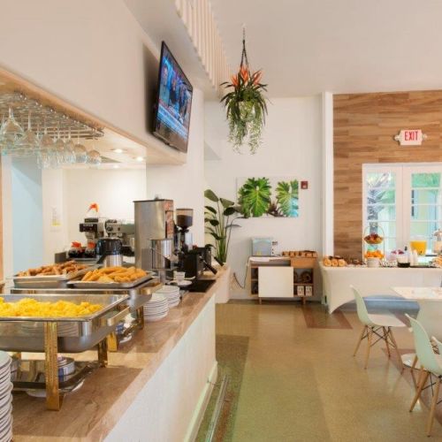 The image shows a well-lit dining area with buffet-style food setup, including trays of food, dining tables and chairs, a bar counter, and TV screen.