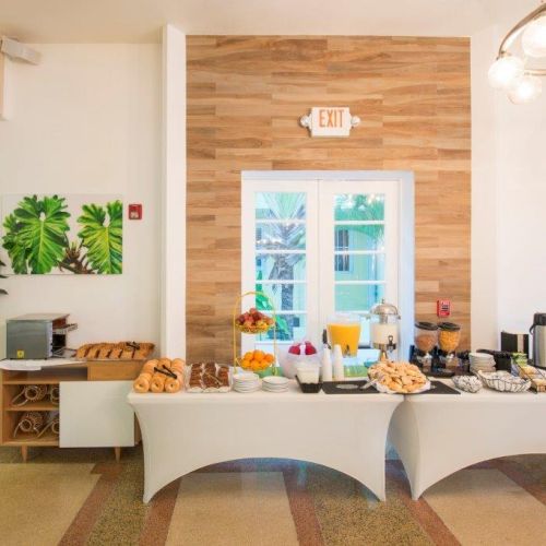 A breakfast buffet setup with various foods, drinks, plants, and a coffee station on white covered tables in a bright room with a door.