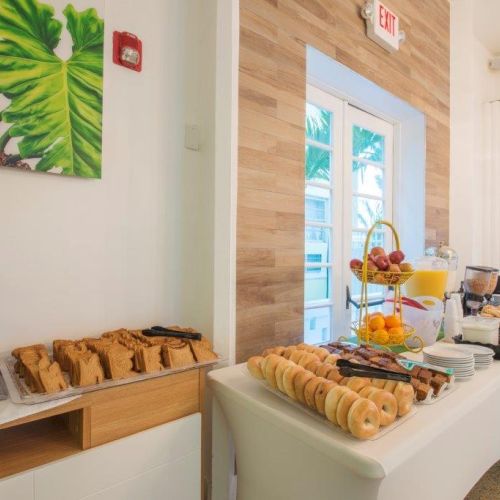 A breakfast buffet table with toast, bagels, fruits, juices, and various breakfast items arranged neatly in a bright, open-room setting.