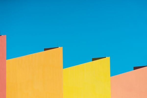 The image shows a row of colorful, angular rooftops in shades of orange, yellow, and pink against a clear, blue sky.