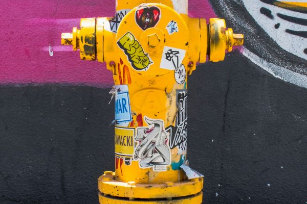 A yellow fire hydrant covered in stickers stands in front of a colorful graffiti wall. The sidewalk underneath shows some shadowing and chalk markings.