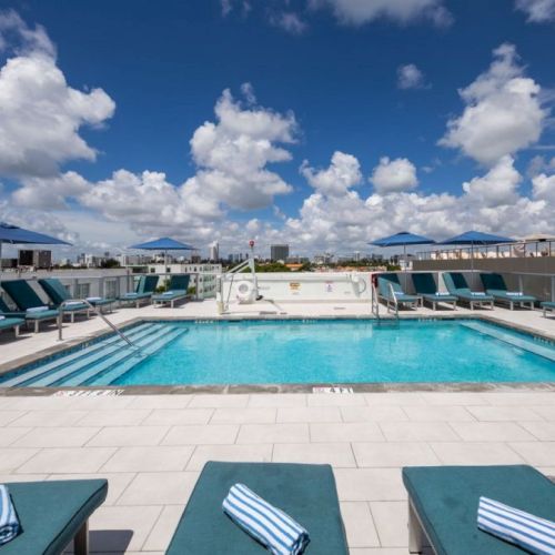 The image shows a rooftop swimming pool with lounge chairs and umbrellas, set against a backdrop of a cloudy blue sky.