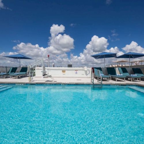 A rooftop swimming pool with clear blue water is surrounded by lounge chairs and umbrellas under a partly cloudy sky.