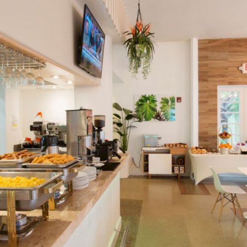 A modern breakfast buffet area with hot food, beverages, chairs, and tables, featuring a bar stocked with liquor bottles and a TV mounted on the wall.