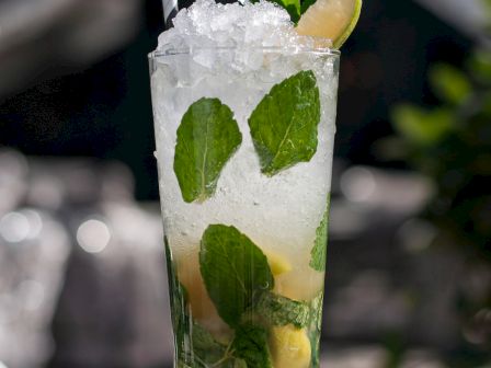 A refreshing Mojito cocktail is served in a clear glass with mint leaves, lime wedges, a straw, and crushed ice, placed outdoors on a table.