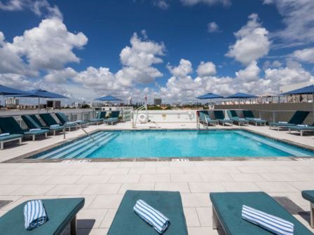 This image shows a rooftop swimming pool with lounge chairs and striped towels under a sky with scattered clouds.