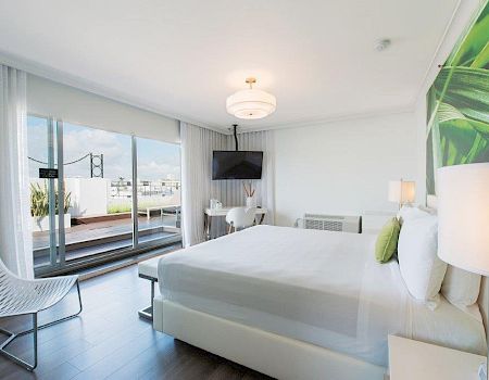 A modern bright bedroom with a large bed, wall TV, sheer curtains, and glass doors to a balcony with outdoor view.