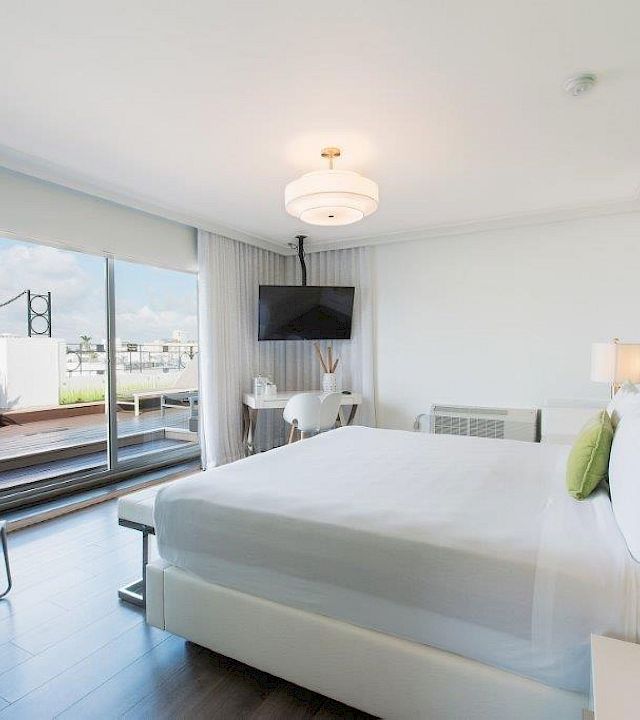 A modern bright bedroom with a large bed, wall TV, sheer curtains, and glass doors to a balcony with outdoor view.