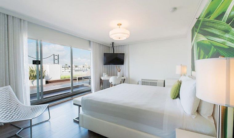 A modern bright bedroom with a large bed, wall TV, sheer curtains, and glass doors to a balcony with outdoor view.
