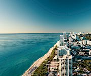 Aerial view of a tropical coastline with a sandy beach, high-rise hotels, and a distant city skyline by the ocean.
