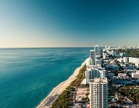 Aerial view of a tropical coastline with a sandy beach, high-rise hotels, and a distant city skyline by the ocean.