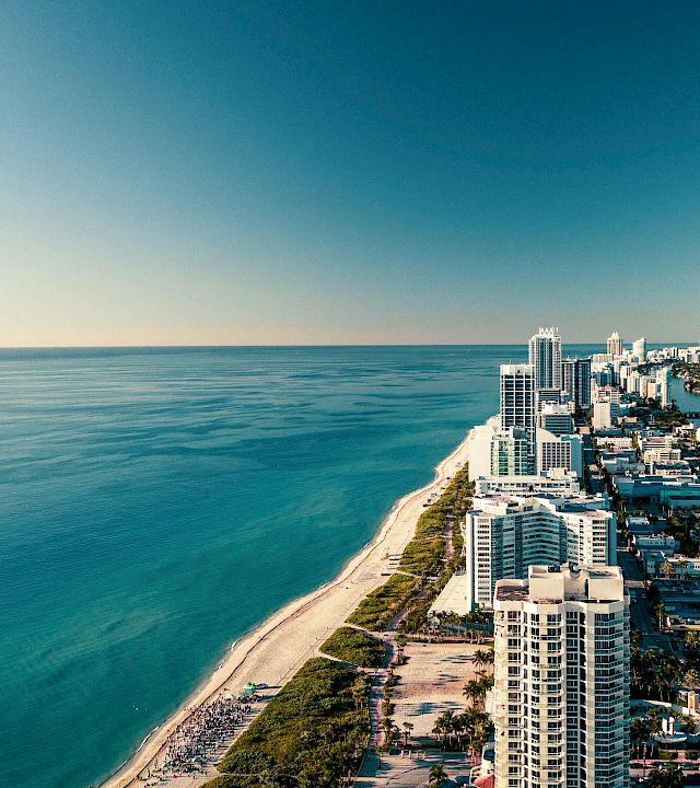 Aerial view of a tropical coastline with a sandy beach, high-rise hotels, and a distant city skyline by the ocean.