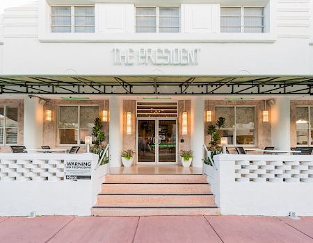 Exterior courtyard of a modern building with patio seating under green awnings, and the sign “THE RESIDENT” above the entrance.