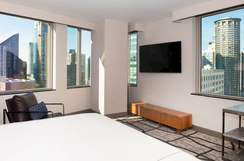 A modern hotel room with large windows showcasing a cityscape, a bed, a TV, a chair, and a small table.