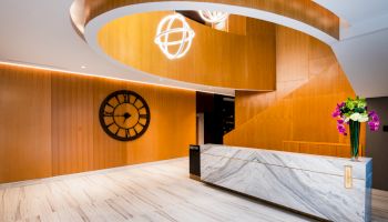 The image shows a modern reception area with a sleek marble counter, a wall clock, vibrant flowers, and a stylish, curved wooden staircase.
