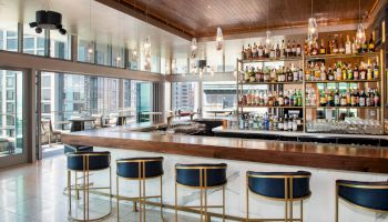 A modern bar with rows of liquor bottles, four blue barstools, large windows, and a wooden ceiling, overlooking an urban cityscape.