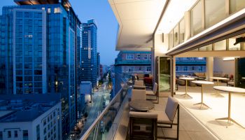 The image shows a modern cityscape at dusk with tall buildings and an elegant, open-air balcony furnished with tables and chairs.