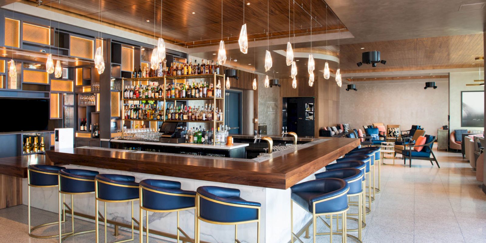 A modern bar with a wooden countertop, blue stools, a well-stocked liquor shelf, hanging pendant lights, and a lounge area in the background.