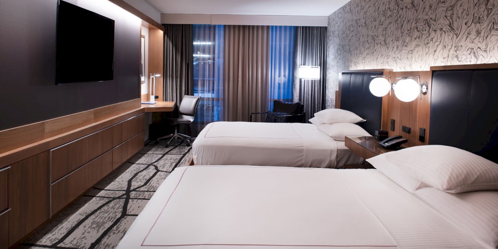 A modern hotel room with two neatly made beds, a flat-screen TV on the wall, a desk and chair near the window, and stylish decor and lighting.