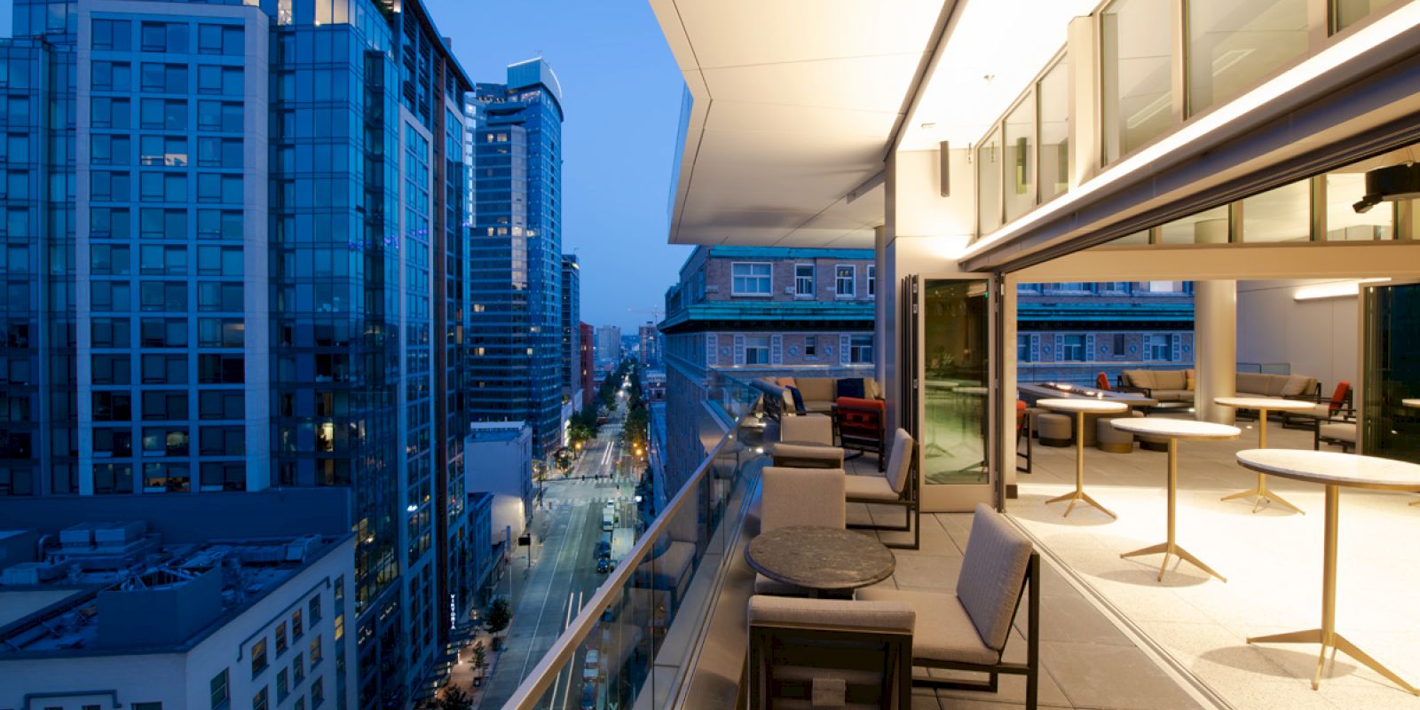 The image shows an outdoor balcony of a modern building with a view of city skyscrapers and a street below, featuring tables and seating.