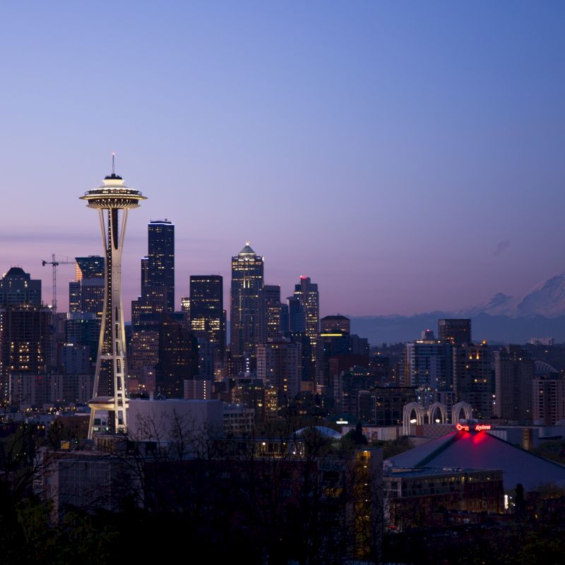 The image shows a city skyline at dusk with prominent tall buildings and a distinctive tower, and a mountain in the background.