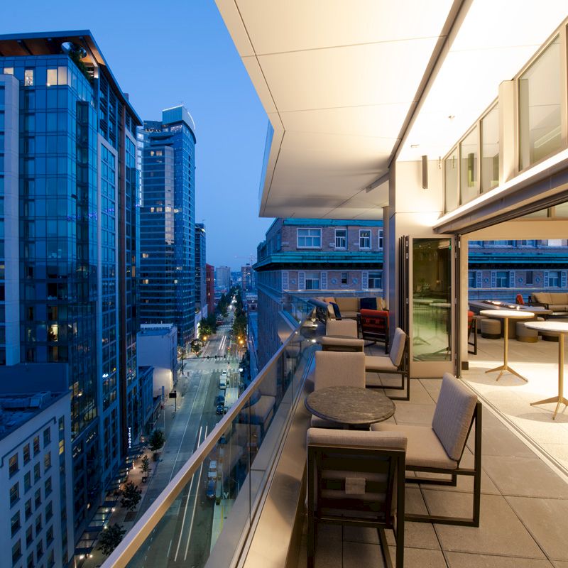 The image shows a modern balcony with tables and chairs overlooking a cityscape of tall buildings during twilight.